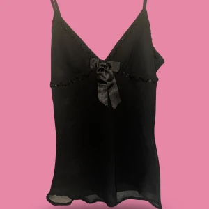 Svart linnetopp med rosett och pärlor - Snygg svart linnetopp med smala axelband och v-ringning. Toppen har dekorativa svarta pärlor längs halsringningen och under bysten, samt en glansig rosett framtill. 