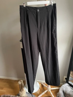 Svarta kostymbyxor med rak passform - Snygga svarta kostymbyxor med rak passform och klassisk knappstängning framtill. Byxorna har en stilren look och är tillverkade i ett mjukt tyg som ger en skön känsla. Perfekta för dig som gillar minimalistisk stil och vill ha ett par byxor som funkar till många outfits.