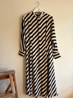 Svartvit långrandig klänning H&M - Säljer en långklänning från H&M i storlek S med svartvita diagonala ränder. Klänningen har lång ärm, rund hals och är tillverkad i mjuk polyester. Den har en loose passform och stängs med knappar framtill och snöre i midjan.  Perfekt för dig som gillar grafiska mönster och stilren design.