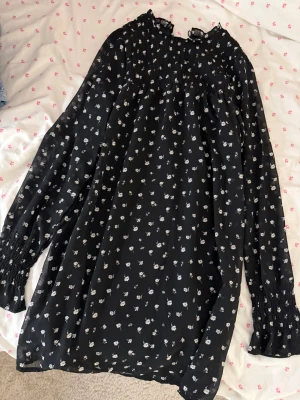Svart blommig klänning - svart kortklänning från H&M i storlek XS. Klänningen har vita små blommor, smock upptill och långa ärmar med smockade muddar. Luftigt material.