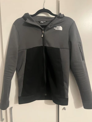 Grå och svart hoodie från The North Face - Snygg hoodie från The North Face i grått och svart med huva och hel dragkedja framtill. Tröjan har långärmade ärmar och en praktisk ficka med dragkedja på ena armen. Perfekt för chill dagar eller när du vill ha en sportig look.