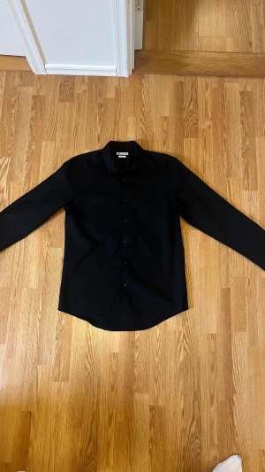 Svart slim fit skjorta från Zapa - Stilren svart skjorta från Zapa i slim fit-modell. Skjortan har klassisk krage, långa ärmar och knäppning framtill. Materialet är mjukt och känns som bomull, perfekt för dig som gillar en clean och enkel look.