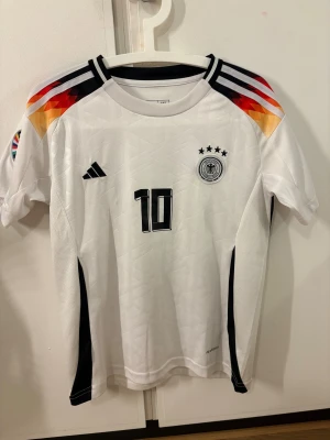 Tysklands Musiala #10 fotbollströja Adidas - Vit Tyskland fotbollströja från Adidas med Musiala och nummer 10 på ryggen. Tröjan har svarta detaljer, mönstrade axlar i rött, orange och gult, samt broderat förbundsmärke med fyra stjärnor. Tillverkad i lätt funktionsmaterial.