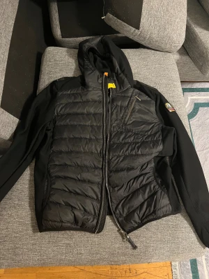 Parajumper Cardigan - Snygg svart pufferjacka från Parajumpers som har aldrig använts, med huva och quiltad framsida. Jackan har dragkedja, sidofickor och en patch på. Jackan har en modern, normal passform. Perfekt för dig som vill ha en stilren look med coola detaljer.