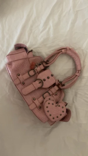 Rosa handväska med hjärt-detalj - Säljer en rosa handväska i croco-mönstrat syntetmaterial med coola metalldetaljer och bälten. Väskan har ett hjärtformat hänge med nitar och ett körsbärs-berlock. Handtaget är vadderat och väskan har flera fack och ringar.