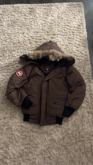 Brun Canada Goose dunjacka med päls - Säljer en brun Canada Goose dunjacka med fet päls på huvan och klassisk patch på ärmen. Jackan har flera fickor, dragkedja och knappar framtill samt ribbade muddar. Perfekt för kalla vinterdagar och har en riktigt schysst look.