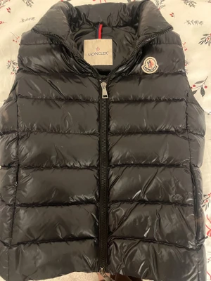 Svart dunväst från Moncler - Svart, glansig dunväst från Moncler med quiltad design och hög krage. Den är använd men forfarande i väldigt bra skick. Storleken Xs eller 0.