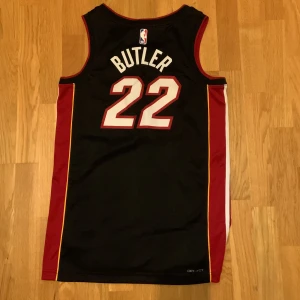 22/23 Miami Heat Butler  - Snygg Miami Heat baskettröja med Butler 22 på ryggen. Svart bas med röda och gula detaljer längs sidorna, vit text och Nike-logga framtill. Tillverkad i lätt och ventilerande Dri-FIT-material, perfekt för träning eller match.