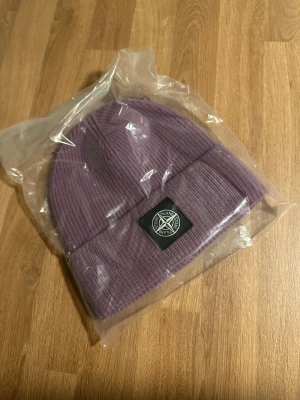 Lila ribbad mössa från Stone Island - Snygg lila mössa från Stone Island med ribbad struktur och klassisk uppvikt kant. Märkets ikoniska logopatch framtill ger en clean och trendig look. Perfekt för att hålla värmen och samtidigt se fresh ut under kyliga dagar.