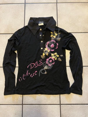 Svart blommig skjorta från D&G - Svart långärmad skjorta från D&G med stor krage och knappar framtill. Framsidan har broderade blommor i rosa och gult samt texten 'D&G chic' i rosa. Skjortan är gjord i mjuk bomull och har en slim passform som sitter snyggt på kroppen.