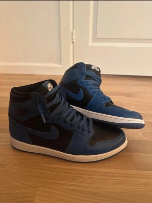 Nike Air Jordan 1 blå och svarta - Säljer ett par Nike Air Jordan 1 sneakers i blått och svart med högt skaft. Skorna har klassisk snörning, rund tå och vit platt sula. Ikonisk Jordan-logga på sidan och Nike Air-märke på plösen. Perfekt för dig som gillar streetwear och snygga färgkombinationer.