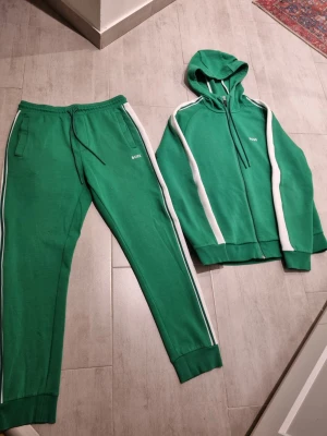 Grön hoodie från BOSS med vita ränder - Snygg grön hoodie från BOSS med vita ränder längs ärmarna och dragkedja framtill. Tröjan har huva med snörning och ribbade muddar. Perfekt för chill eller träning, med sportig look och bekväm passform.