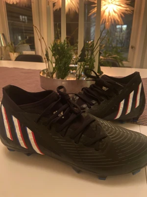 Svarta adidas fotbollsskor med blå dobbar - Säljer ett par svarta adidas fotbollsskor med snörning och platt sula med blå dobbar. Skorna har de klassiska adidas-ränderna i vitt, rött och blått på sidan. Ovandelen är i syntetmaterial och har en rund tå. Perfekt för dig som vill ha snygga och funktionella skor på planen.