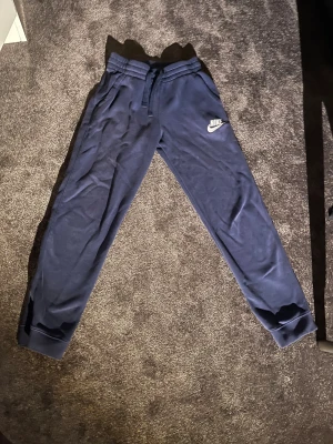 Mörkblå mjukisbyxor från Nike - Storlek 147-158. Snygga mörkblå mjukisbyxor från Nike med broderad logga på vänster ben. Byxorna har resår och snörning i midjan samt ribbade muddar vid bensluten. Tillverkade i mjukt och skönt material som passar perfekt för chill eller träning.