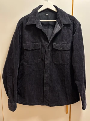 Svart manchester overshirt från Uniqlo - Säljer en svart overshirt från Uniqlo i mjuk manchester. Jackan har klassisk krage, två bröstfickor med knappar och raka ärmar. Perfekt lager-på-lager-plagg med avslappnad passform och snygg struktur.