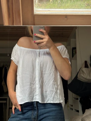 Vit blus - Säljer en luftig vit blus med korta ärmar och knappar framtill. Blusen har en rundad halsringning som kan bäras offshoulder och är dekorerad med små spetsdetaljer vid ärmsluten. Materialet är tunt och skönt, perfekt att ha på stranden en varm dag🩷