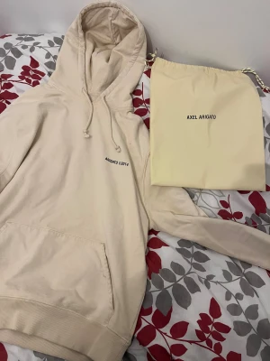 Beige hoodie Axel Arigato - Säljer en beige hoodie från Axel Arigato med svart tryck både fram och bak. Tröjan har huva med snörning, stor magficka och ribbade muddar. Materialet är mjuk bomull och passformen är normal. Perfekt för dig som gillar stilrena streetwear-plagg.