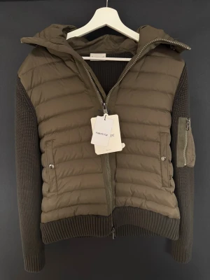 Moncler mörkgrön pufferjacka med huva - Snygg pufferjacka från Moncler i mörkgrön färg med stickade ärmar och detaljer. Jackan har huva, två sidofickor med knappar, dragkedja framtill och en extra ficka med dragkedja på ärmen. Perfekt för dig som gillar exklusiva märken och cool design. Använd enbart en gång. Priset är inte hugget i sten!