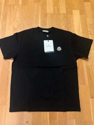 Svart Moncler t-shirt i bomull - Svart t-shirt från Moncler i mjuk bomull med klassisk rund hals och korta ärmar. På bröstet sitter Monclers ikoniska logopatch i vitt och rött. Enkel och stilren design som passar perfekt till jeans eller shorts.