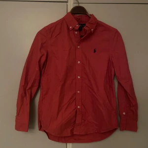 Ralph Lauren Skjorta - Riktigt snygg röd skjorta från Ralph Lauren med klassisk button-down krage och ikoniska broderade loggan på bröstet. Skjortan har långa ärmar, vita knappar framtill och är gjord i mjuk bomull. Jag är 170 lång och väger 57 kg, den passar perfekt på mig! Perfekt för dig som gillar stilrena och tidlösa plagg.