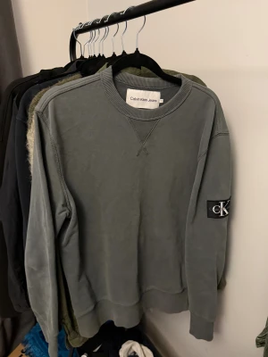 Olivgrön sweatshirt från Calvin Klein Jeans - Snygg sweatshirt från Calvin Klein Jeans i storlek S. Tröjan har rund hals, ribbade muddar och en svart patch med CK-logga på vänster ärm. Perfekt för dig som gillar stilrena och klassiska plagg med en modern touch.