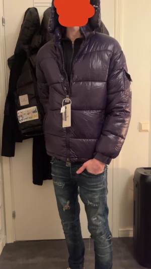 Mörkblå Moncler pufferjacka med huva - Säljer en mörklila pufferjacka från Moncler med huva och dragkedja framtill. Jackan har en ficka med knapp på ärmen och Moncler-logga. Glansigt material och quiltad design, perfekt för kalla dagar. Märkesetikett och autenticitetsbricka medföljer.