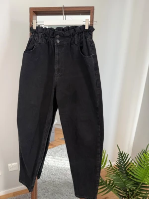 Svarta paperbag jeans från Vila - Snygga svarta paperbag jeans från Vila med hög midja och rynkad kant upptill. Modellen har raka ben, klassiska fickor fram och bak samt knapp och dragkedja. Perfekt för dig som gillar en chill och trendig look.