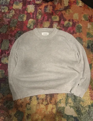 Grå stickad tröja från Maison Margiela - Säljer en grå stickad tröja från Maison Margiela med klassisk rund hals och ribbad struktur. Tröjan har långa ärmar och de ikoniska vita stygn-detaljerna i nacken. Perfekt för dig som gillar minimalistisk och clean stil.