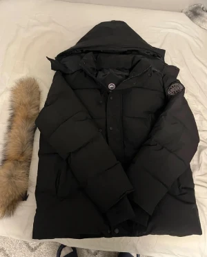 Svart dunjacka från Canada Goose - Säljer en svart dunjacka från Canada Goose med avtagbar huva och pälskant. Jackan har klassisk patch på ärmen, stora fickor och stängs med både dragkedja och knappar. Perfekt för kalla vinterdagar och riktigt snygg streetstil.