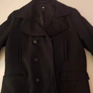Mörkblå kappa - Snygg svart kappa från H&M med quiltat foder och knappar framtill. Perfekt för kyligare dagar med sin stilrena design och praktiska fickor. Ett måste för garderoben! Pris ej hugget I sten 
