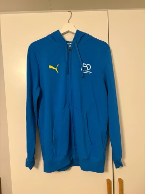 Blå Puma hoodie Gothia Cup M - Blå hoodie från Puma i storlek M med Gothia Cup 50-års tryck på bröstet och gul Puma-logga. Tröjan har huva med snörning, dragkedja framtill och två stora fickor. Perfekt för dig som gillar sportig stil och vill sticka ut med en unik Gothia Cup edition.