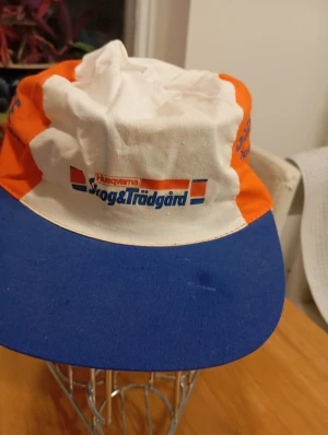 Husqvarna keps Skog & Trädgård - Säljer en retro keps från Husqvarna med Skog & Trädgård-tryck. Kepsen har vit och orange krona med blå skärm, samt blå och orange detaljer. Perfekt för dig som gillar klassiska arbetskläder med cool logga. Materialet är bomull.