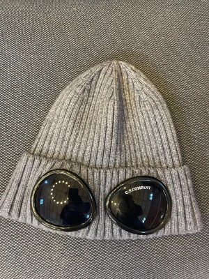 Grå C.P. Company Goggle Beanie - Säljer en grå ribbstickad mössa från C.P. Company med ikoniska svarta goggle-linser framtill. Mössan är tillverkad i mjuk ull och har en uppvikt kant. Perfekt för dig som vill sticka ut med streetwear-vibe och snygga detaljer.