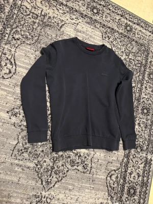 Mörkblå sweatshirt från Hugo Boss - Snygg mörkblå sweatshirt från Hugo Boss med diskret broderad HUGO-logga på bröstet. Tröjan har rund hals, långa ärmar och ribbade muddar vid ärmslut och nederkant. Perfekt för en chill och stilren look.