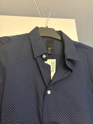 Mörkblå prickig skjorta - Mörkblå skjorta från H&M i slim fit-modell med små vita prickar över hela tyget. Skjortan har klassisk krage, långa ärmar och vita knappar framtill. Perfekt för dig som gillar stilrena detaljer och en modern passform.