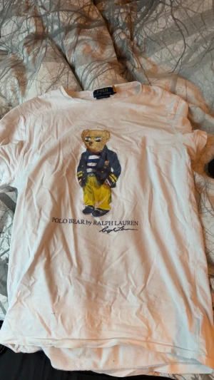 Vit Polo Bear t-shirt från Ralph Lauren - Vit t-shirt från Polo Ralph Lauren med ikoniskt Polo Bear-tryck på framsidan. Modellen är custom slim fit och har korta ärmar. Materialet är mjuk bomull och trycket visar en nalle i gul byxa och blå kavaj. Perfekt för dig som gillar klassiska street vibes. Strl S passar xs 