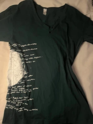 Mörkgrön t-shirt med vit textprint - Snygg mörkgrön t-shirt med v-ringad hals och korta ärmar. Framsidan har en cool vit textprint som ger en unik och personlig vibe. Perfekt för dig som gillar streetstyle och vill sticka ut lite extra. 