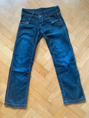 Mörkblå vaxade jeans med knappar och fickor - Unika mörkblå jeans med kontrastsömmar och coola knappar på både fram- och bakfickor. Byxorna har raka ben och bred linning med extra detaljer. Perfekta för dig som vill sticka ut med en egen stil. Kostar vanligtvis 2000 men säljer dem för det jag köpte dem för. Skulle inte säga att dem sitter skinnet utan loose.