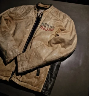 Beige vintage läderjacka  - Snygg beige skinnjacka från Jack & Jones Vintage Denim med svarta detaljer längs sidorna och dragkedja framtill. Jackan har ståkrage med tryckknappar och tryckt text på bröstet. Otrolig jacka i bra kvalite! 