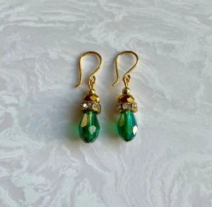 Gröna droppformade örhängen med guld - Snygga örhängen med droppformade, facetterade gröna pärlor och guldfärgade detaljer. Mellan pärlorna sitter en glittrig ring med små stenar som ger extra bling. Krokarna är också guldfärgade och hela designen känns både elegant och trendig.