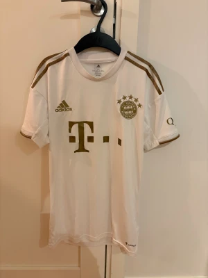 Vit/guldig FC Bayern München tröja - Snygg vit/guldig FC Bayern München fotbollströja från Adidas i storlek XS. Tröjan har guldiga detaljer, logga och tryck, samt klassiska Adidas-ränder på axlarna. Tillverkad i lätt och ventilerande polyester, perfekt för träning eller match. Denna tröjan går inte att få tag på längre, den var limiterad. Detta är en äkta matchtröja köpt på en officell adidas butik.