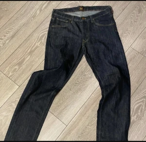 Mörkblå Lee jeans med raka ben - Snygga mörkblå jeans från Lee med klassisk femficksdesign och raka ben. Jeansen har kontrastsömmar, Lee-logga på läderpatch baktill och knäppning med knappar framtill. Perfekt för dig som gillar tidlös stil och kvalitetsjeans.