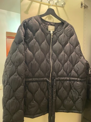 Svart quiltad pufferjacka från H&M - Quiltad svart pufferjacka från H&M i storlek XXL. Jackan har rund hals, dragkedja framtill och ett bälte i midjan för snygg passform. Materialet är syntet och jackan har ett coolt mönster med droppformade quiltningar. Perfekt för höst och vinter.