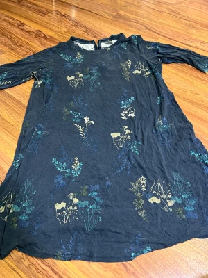 Blommig marinblå klänning Lindex 2XL - Snygg marinblå klänning från Lindex i storlek 2XL med blommigt mönster i blått, beige och grönt. Klänningen har lång ärm, rund hals och dragkedja i ryggen. Mjuk bomullskänsla och avslappnad passform, perfekt för dig som gillar stilrena och bekväma plagg.