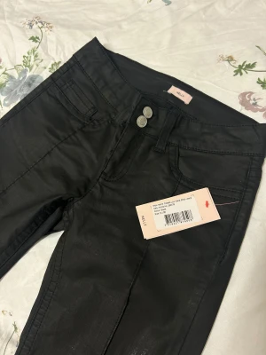 Svarta bootcut jeans från Nelly, strl 36 - Snygga svarta jeans från Nelly i bootcut-modell med låg midja. Jeansen har två framfickor, dubbla knappar i midjan och coola broderade detaljer på bakfickorna. Materialet har en lätt glansig finish som ger en trendig look.