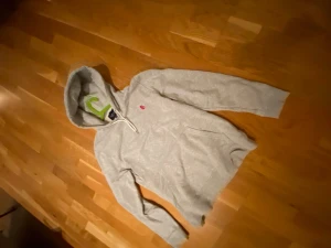 Grå hoodie från Bondelid med huva - Snygg grå hoodie från Bondelid med vit snörning i huvan och röd broderad logga på bröstet. Tröjan har en stor ficka framtill och långa ärmar med ribbade muddar. Perfekt för chill dagar och enkel att matcha med jeans eller joggers.
