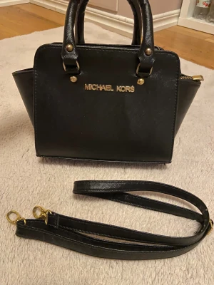 Svart handväska - Snygg svart handväska från Michael Kors med guldfärgade detaljer och logotyp framtill. Väskan har både handtag och avtagbar axelrem, samt dragkedja upptill. Insidan är fodrad med MK mönster ‼️vet ej om den är äkta‼️
