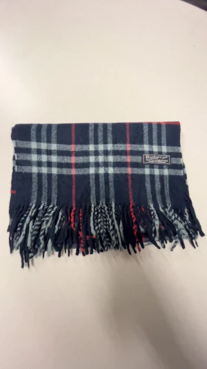 Rutig mörkblå halsduk från Burberrys - Snygg halsduk från Burberrys i klassiskt rutig design med mörkblå, grå och röda toner. Gjord av 100% lammull och har stiliga fransar i ändarna. Perfekt för att ge din outfit en lyxig touch under kyliga dagar.