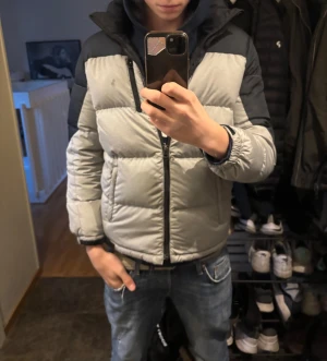 Grå och svart pufferjacka Polo Ralph Lauren - Säljer en grå och svart pufferjacka från Polo Ralph Lauren Performance. Jackan har huva, dragkedja framtill och två sidofickor med dragkedja. Fylld med syntetmaterial och har elastiska muddar vid ärmslut. Perfekt för kalla dagar och riktigt snygg streetstil. Han på bilden är runt 176.