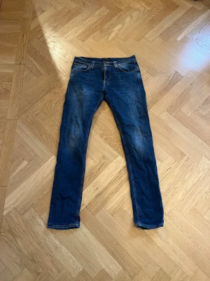 Blå jeans från Nudie Jeans Co - Snygga blå jeans från Nudie Jeans Co med klassisk femficksdesign och orangea sömmar på bakfickorna. Jeansen har slim passform och normal midja, tillverkade i slitstarkt denim. Perfekta för dig som gillar stilrena och tidlösa jeans. Hör av er vid frågor